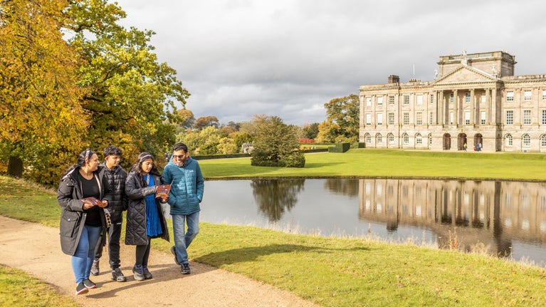 Visitors exploring the garden on a family trail, with a lake and the house in the background in Autumn at Lyme, Cheshire